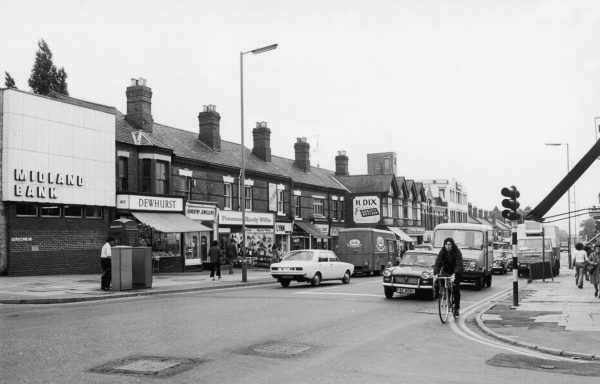 A busy street of shops in the 1970s with cars and people