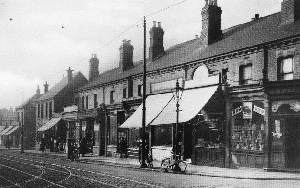 A street of shops in early 20th century, with tramlines and bikes